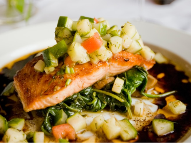 Grilled salmon on top of cooked greens with cucumber and tomato salad with sauce on a white plate